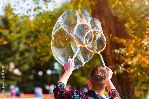 An animator launches large soap bubbles at a children's party using two rings. Autumn nature on a sunny day. The shape of rainbow balls is similar to a heart.  photo