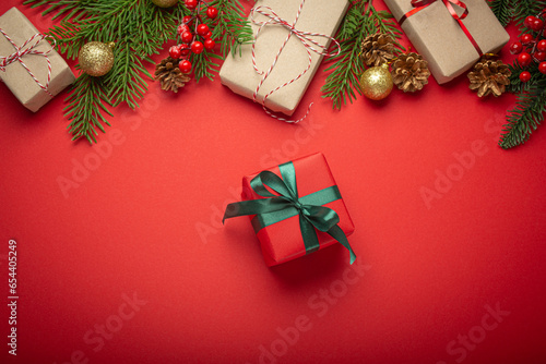 Christmas or New Year celebration red paper festive background with decoration fir tree, present boxes, cones, berries, sparkly red balls and a red wrapped gift in center.