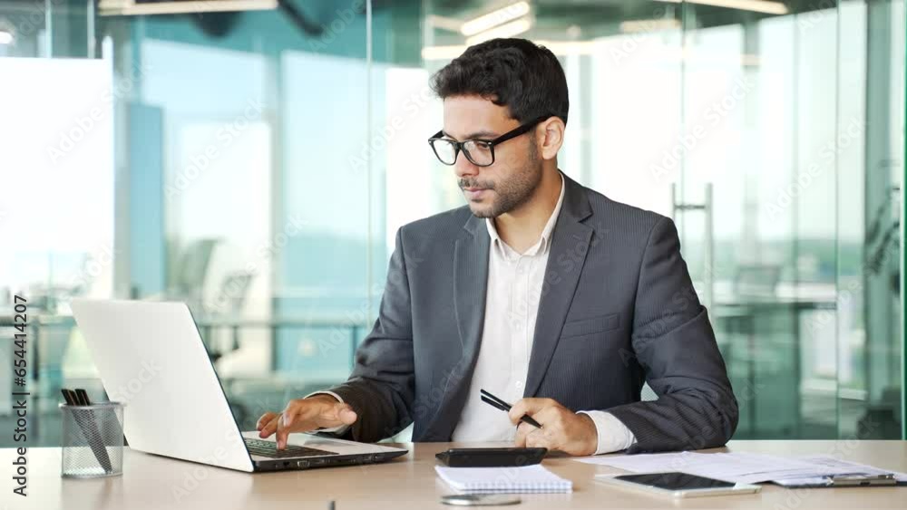 Young financier in a formal suit making financial calculations using a calculator, typing on a laptop, taking notes sitting at workplace. Confident businessman working on startup in business office Young financier in a formal suit making financial calculations using a calculator, typing on a laptop, taking notes sitting at workplace. Confident businessman working on startup in business office