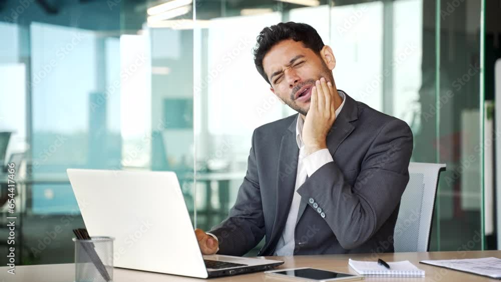 Sad businessman in a formal suit suffering from toothache working on laptop sitting at workplace at desk in business office. Young entrepreneur holds his hand on his cheek, massaging the sore spot Sad businessman in a formal suit suffering from toothache working on laptop sitting at workplace at desk in business office. Young entrepreneur holds his hand on his cheek, massaging the sore spot