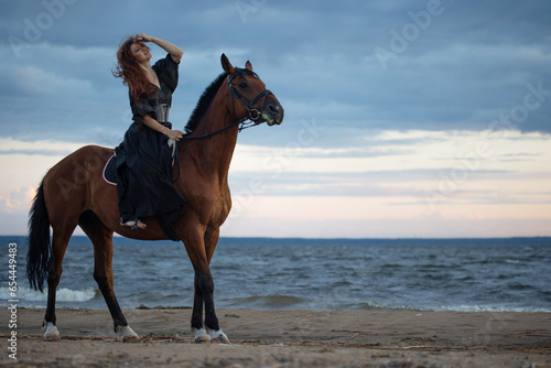 A beautiful young horsewoman in a black dress and with her hair down  riding a horse  portrait against the background of the evening sky  horseback riding in the open air