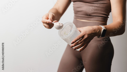 Athletic woman in sportswear with measuring spoon in her hand puts a portion of whey protein powder into a shaker, making protein drink, cropped image