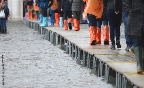 people in gaiters boots walking over elevated walkway during high tide Venice Italy