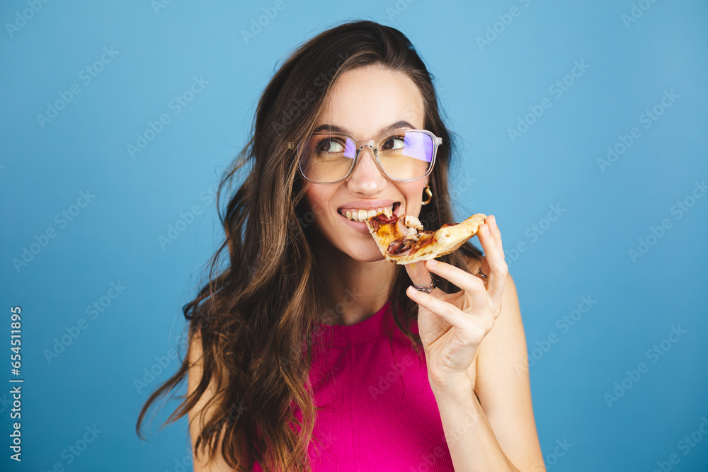 Woman eats slice of pizza, dressed in pink top, isolated blue background. Pretty girl has snack with fast food. Pizza and delivery concept. Happy woman posing with pizza slice, look happy, bite pizza.