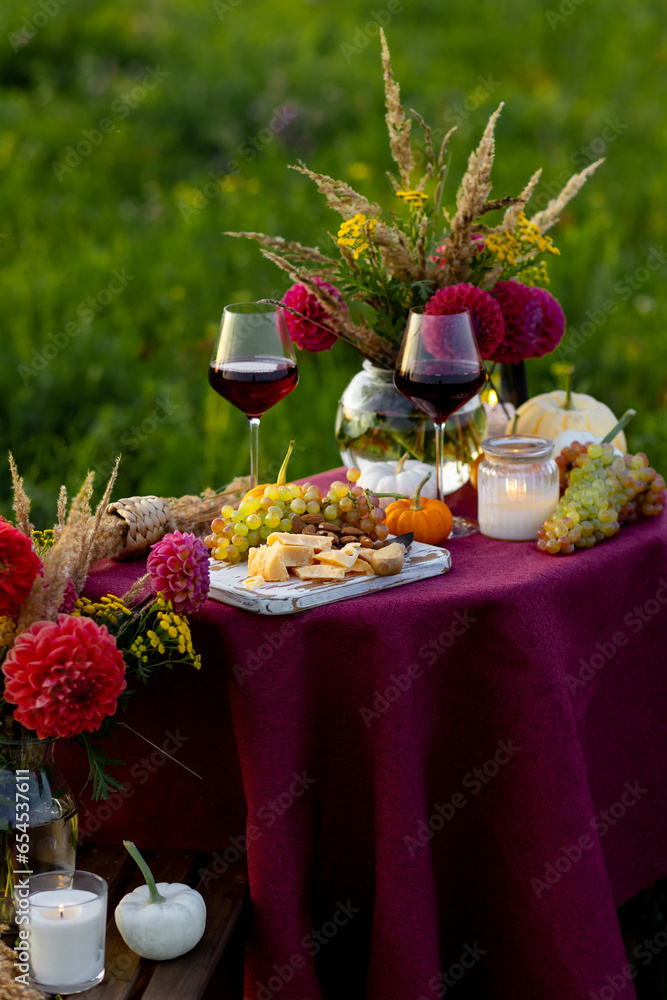 Beautiful decorated candlelight fall dinner, covered with a red tablecloth with flowers, pumpkins, candles, wine glasses. Outdoor in field near the forest in nature. Wedding banquet, sunset, evening