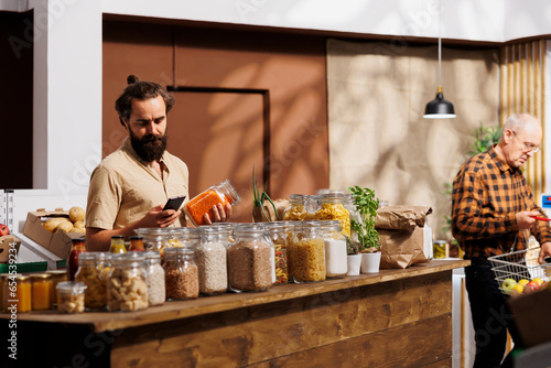 Man in zero waste store analyzing bulk products, using phone to make sure they are freshly harvested. Sustainable living customer thoroughly checking local supermarket food items are additives free