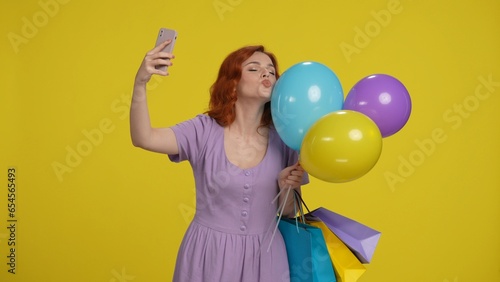 Redhaired woman with shopping bags and colorful balloons takes a selfie on a smartphone. Woman posing kissing balloons on a yellow background.