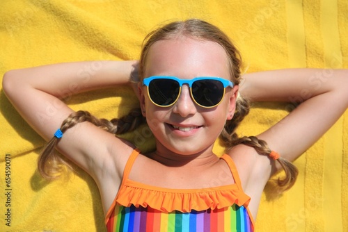 Happy little girl in sunglasses lying on beach towel outdoors, top view