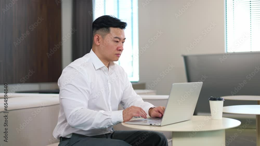 Ambitious asian director with serious facial expression leading business correspondence with foreign partners via remote laptop. Formally dressed man sitting at round desktop and chatting online. Ambitious asian director with serious facial expression leading business correspondence with foreign partners via remote laptop. Formally dressed man sitting at round desktop and chatting online.