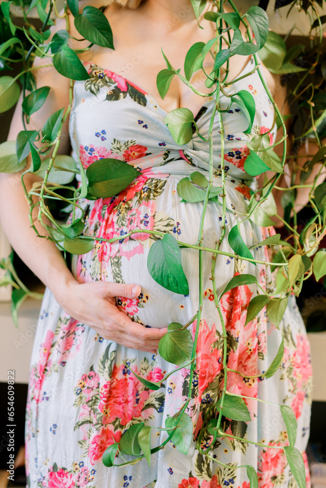 A pregnant woman covers herself with vining plants while wearing a floral dress.