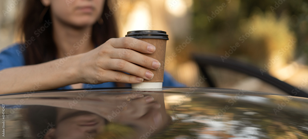 Woman putting coffee takeaway in a paper cup on top of the car roof in the morning, selective focus. Banner.