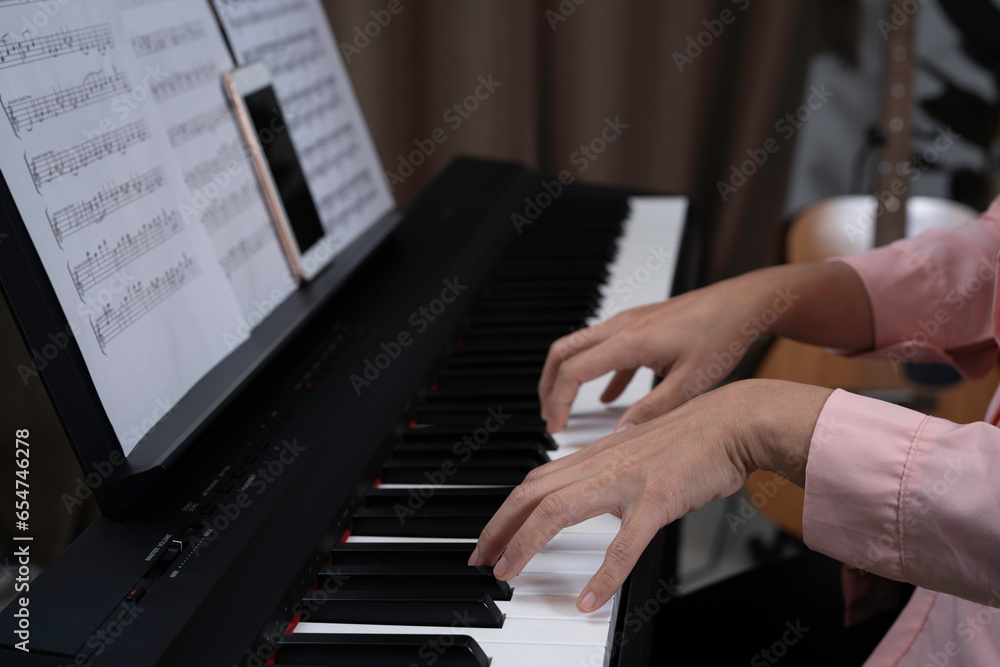 Obraz premium Close up woman hand playing piano at home. Relaxing time concept. Practicing music at home, Note and smartphone put on paino holder. Hands put on keyborad.