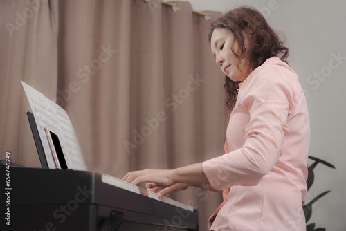 An asian senior woman playing paino with happiness, musician professional in pink shirt lloking at paino keyboard with smartphone and note at note holder. photo