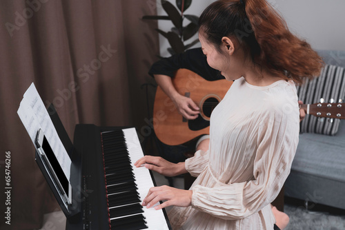 A woman in white dress playing paino with happyness with his friend who playing guitar. Musician practicing paino.