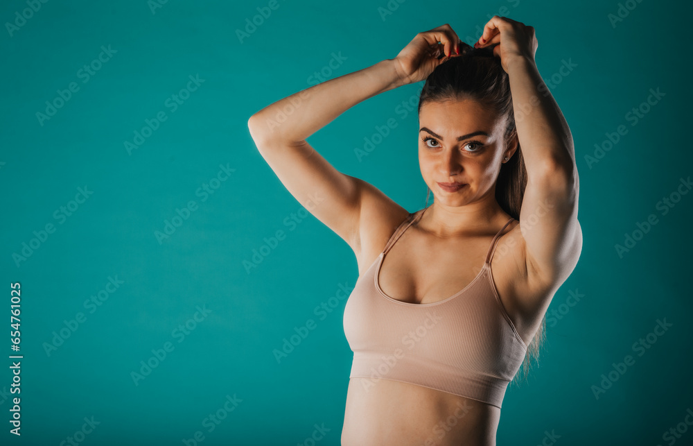 Fototapeta premium A confident fit woman showcases her transformation - muscular arms, defined abs. Inspiring motivation on turquoise background.