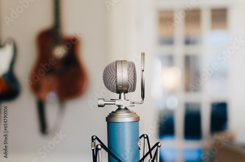 A small bluebird microphone on a stand with a pop filter in a white, bright room with hanging guitars.