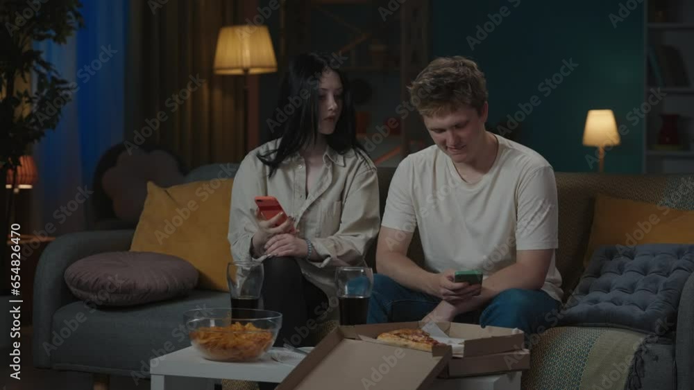 Portrait of teenage couple spending time. Boy and girl sitting on the sofa with smartphones, girl asks something and couple start an argument. Portrait of teenage couple spending time. Boy and girl sitting on the sofa with smartphones, girl asks something and couple start an argument.