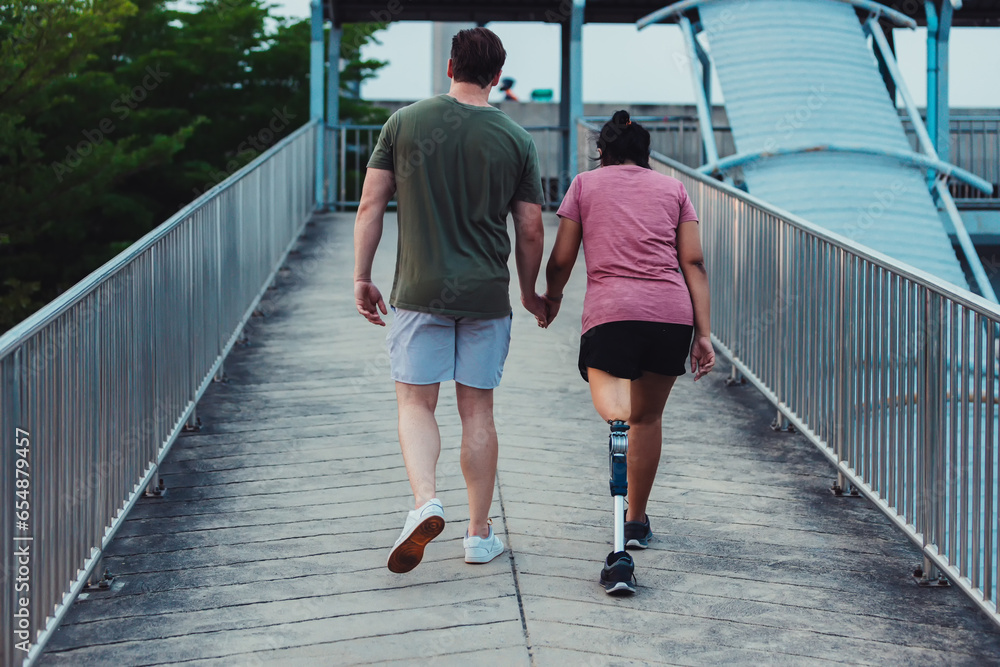 Friend support Friend with a prosthetic leg while exercising outdoor. People walking together on park outdoor. Exercise walking woman with prosthetic leg and friend support together in park outdoor Friend support Friend with a prosthetic leg while exercising outdoor. People walking together on park outdoor. Exercise walking woman with prosthetic leg and friend support together in park outdoor