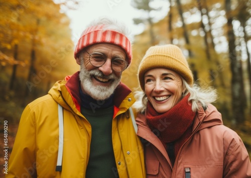 A beautiful portrait of two people standing in a wooded forest wearing autumnal outerwear, with hats and scarves, smiles beaming brightly as they embrace the season