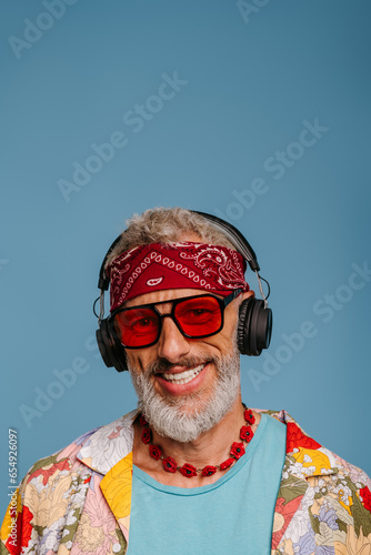 Joyful senior man in funky shirt and headphones listening to the music against blue background