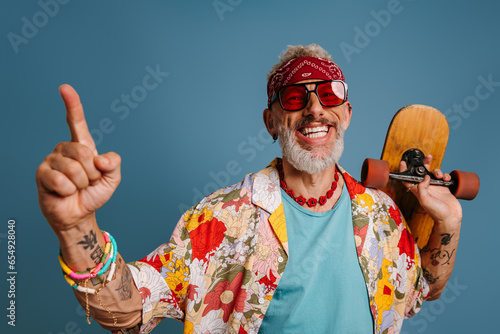 Joyful hipster mature man in stylish funky shirt carrying longboard and gesturing against blue background
