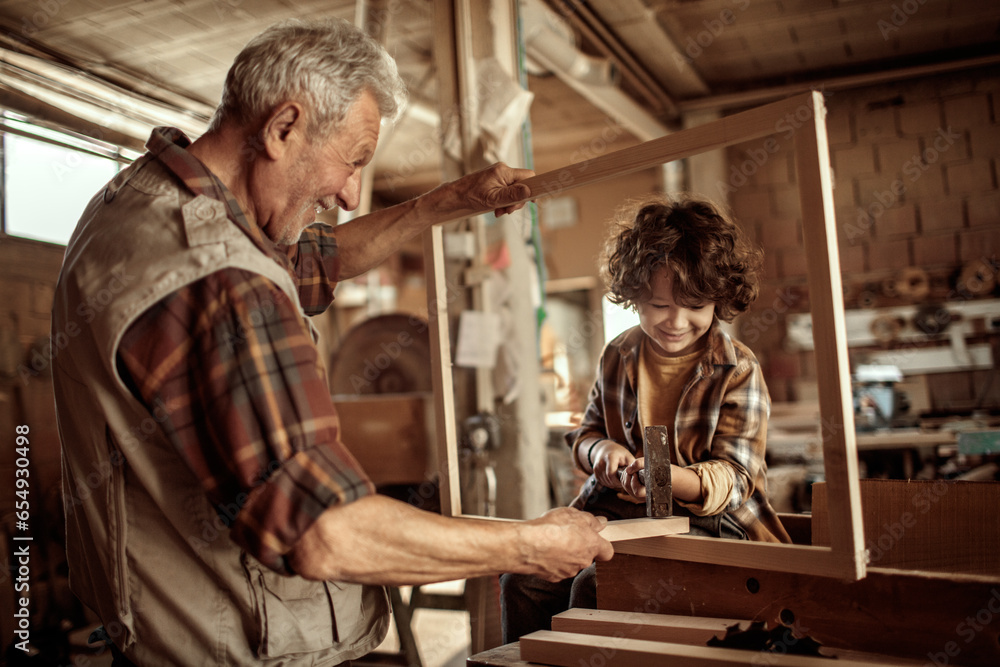 Senior male carpenter teaching his grandson how to use a hammer in a carpentry shop Senior male carpenter teaching his grandson how to use a hammer in a carpentry shop