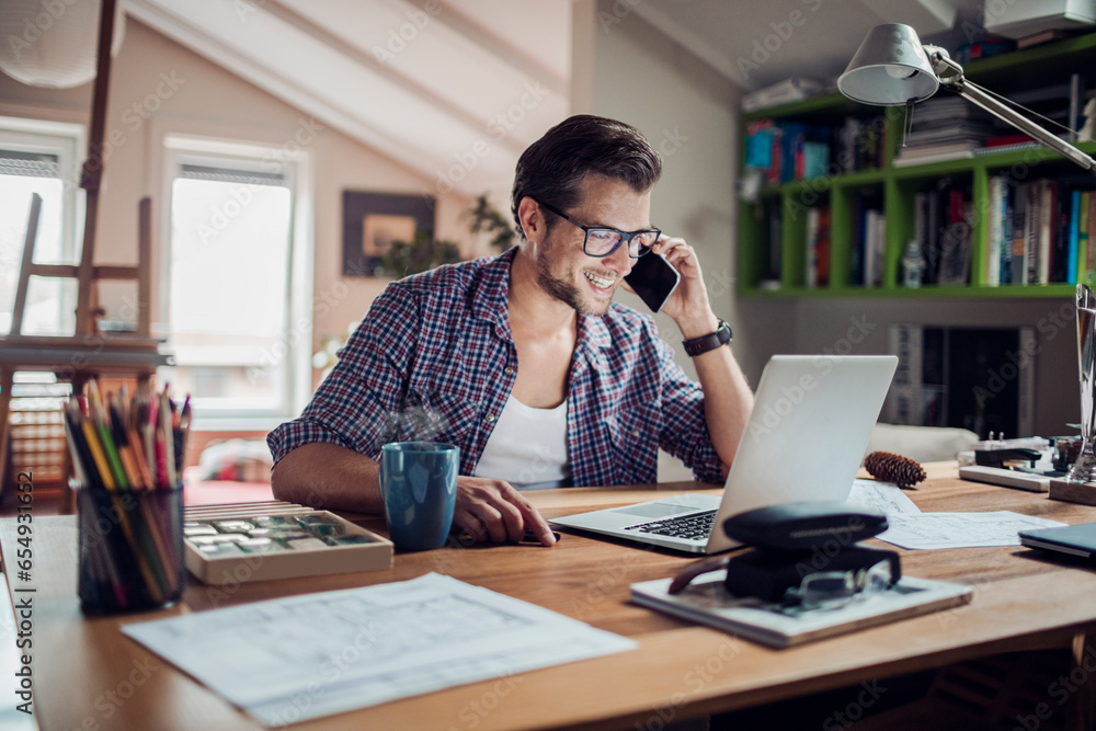Fototapeta premium Young male architect having a conversation on the smartphone while working on design from home