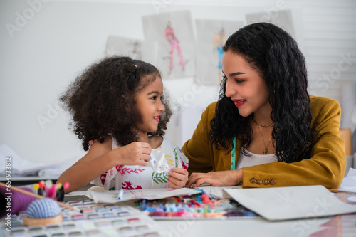 Happy African young mom and her cute daughter learn and teach sewing and design together in the fashion studio.
