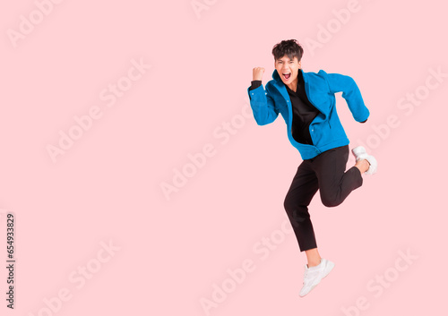 Portrait of happy handsome Asian man in a blue jacket excited and celebrating by jumping up in the air with winner gesture isolated on pink background.