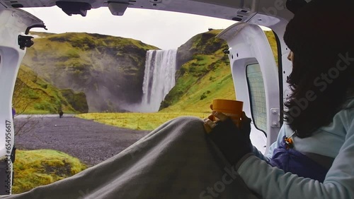 Attractive young female drinking coffee and sitting with blanket inside her campervan while enjoying scenic Skogafoss waterfall, Iceland. Road trip ring road visit Icelandic nature photo