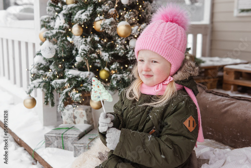 A little girl in winter clothes is sitting on the porch of a house decorated for the Christmas holidays. Christmas holidays for a child.