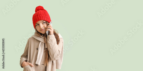 Cute little girl in winter clothes talking by mobile phone on light background with space for text