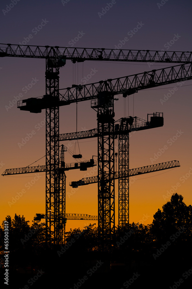 Naklejka premium Silueta de conjunto de torres de grúas al atardecer en un terreno donde se están realizando obras de construcción de edificios residenciales.