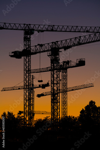Silueta de conjunto de torres de grúas al atardecer en un terreno donde se están realizando obras de construcción de edificios residenciales.