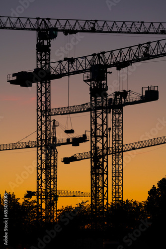 Silueta de conjunto de torres de grúas al atardecer en un terreno donde se están realizando obras de construcción de edificios residenciales.