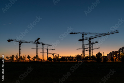Silueta de conjunto de torres de grúas al atardecer en un terreno donde se están realizando obras de construcción de edificios residenciales.