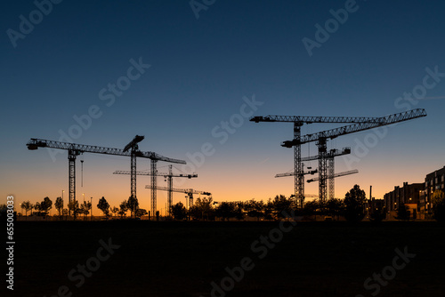 Silueta de conjunto de torres de grúas al atardecer en un terreno donde se están realizando obras de construcción de edificios residenciales.