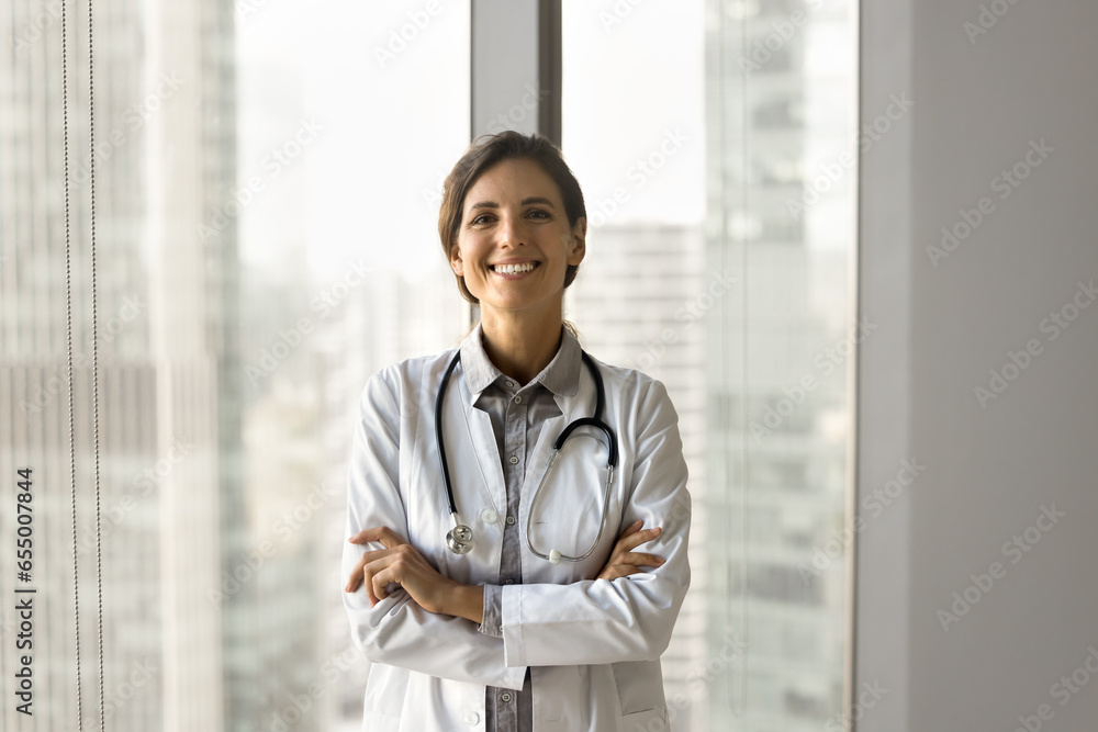 Happy beautiful Latin doctor woman posing at large office window, standing with arms folded, looking at camera with toothy smile. Positive confident medical professional with stethoscope portrait Happy beautiful Latin doctor woman posing at large office window, standing with arms folded, looking at camera with toothy smile. Positive confident medical professional with stethoscope portrait