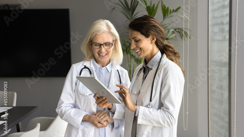 Two positive young and senior doctor women using tablet together, discussing medical application, meeting in boardroom, talking, smiling. Younger practitioner consulting elder colleague