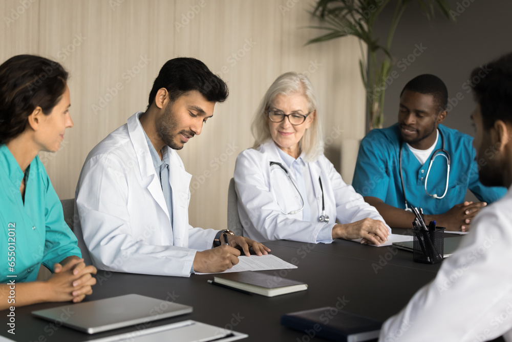 Diverse team of medical professionals making business decision on meeting with colleagues, partners. Confident young Indian chief doctor signing agreement, hiring new specialist Diverse team of medical professionals making business decision on meeting with colleagues, partners. Confident young Indian chief doctor signing agreement, hiring new specialist