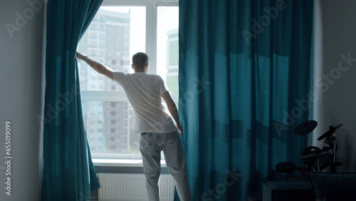 A man in a dark apartment with curtains. Media. An adult in summer clothes is a man who goes to open turquoise curtains.