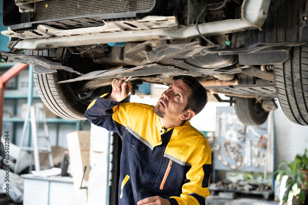 Vehicle mechanic conduct car inspection from beneath lifted vehicle. Automotive service technician in uniform carefully diagnosing and checking car's axles and undercarriage components. Oxus - obrazy, fototapety, plakaty 