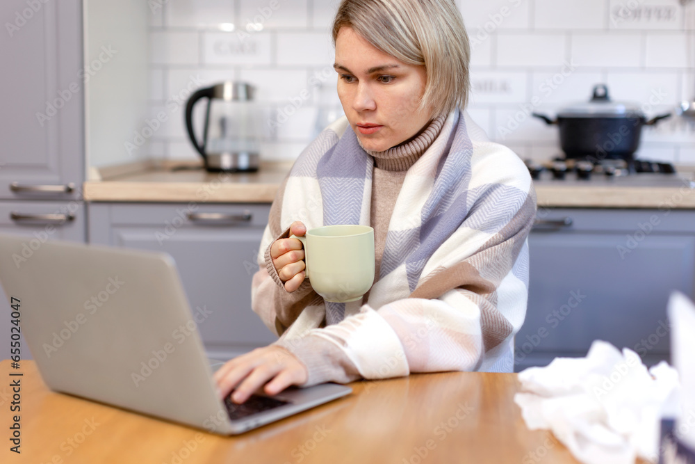 A woman wrapped in a scarf works on a laptop and warms herself with hot tea. A woman works at home and drinks a hot medicinal drink. Sick, work, treatment. A woman wrapped in a scarf works on a laptop and warms herself with hot tea. A woman works at home and drinks a hot medicinal drink. Sick, work, treatment.