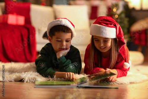 Cute little children in Santa hats reading story at home on Christmas eve