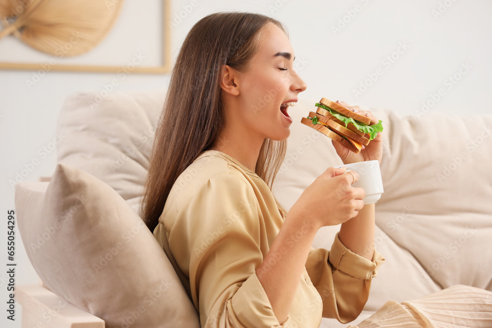 Beautiful young woman with cup of coffee eating tasty sandwich while resting on sofa in living room Beautiful young woman with cup of coffee eating tasty sandwich while resting on sofa in living room