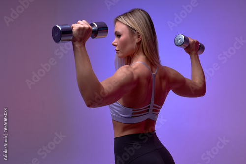 girl demonstrating biceps muscles. Rear view of young woman in modern light beige close-fitting sportswear holding dumbbells and showing fit body isolated purple background.