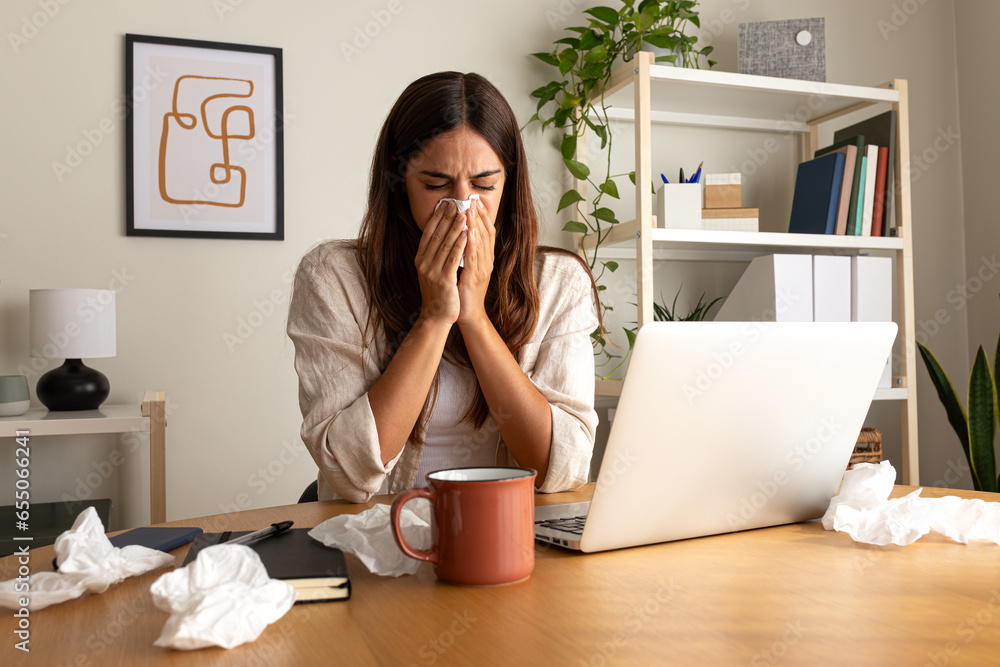 Sick woman working from home office. Caucasian female blowing nose with tissue while working with laptop. - obrazy, fototapety, plakaty 