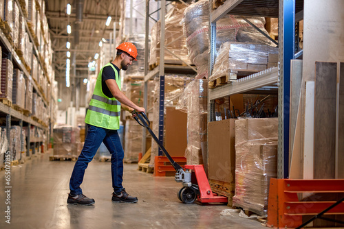 male staff worker in warehouse uses hand pallet stacker to transport goods, alone, dressed in working clothes and safety hard hat.Skilled warehouse employee pushing manual pallet jack