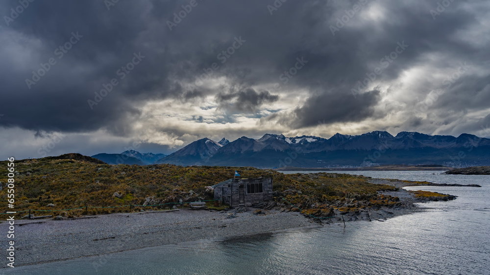 On a small island Bridges in the Beagle Channel, an old dilapidated missionary hut is visible. The flag of Argentina on the roof. Stunted vegetation around. Mountains against a cloudy sky. 