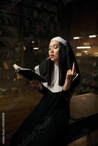 Nun woman reading bible, smoking cigarette and showing obscene hand gesture photo